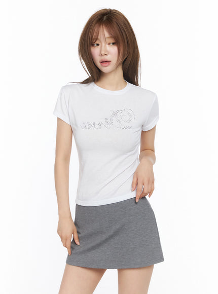 Diva Short Sleeve Tee IU525 White front view on model with slight angle, rhinestone Diva script visible on chest, fitted silhouette with gray mini skirt