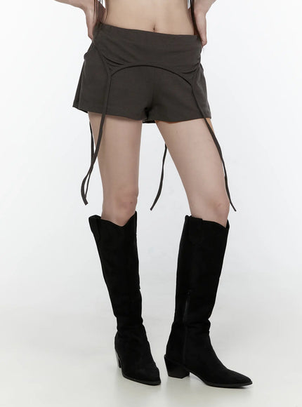 Drawstring Banded Shorts IL522 Dark gray close-up front view on model showing curved arc seam detail, low-rise waist, and multiple hanging straps with cowboy boots