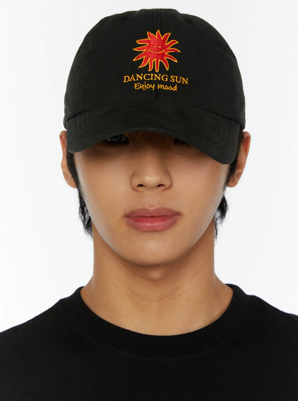 Embroidered Sun Logo Cap IL515 Black close-up front on model face partially hidden by brim, Dancing Sun embroidery detail visible on black cotton