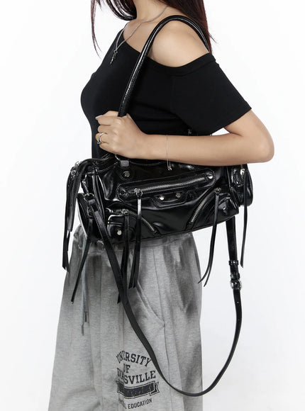Faux Leather Shoulder Bag CL527 Black side view on model shoulder-carried with crossbody strap, glossy faux leather showing multiple zippers and fringe tassels, styled with grey sweatpants