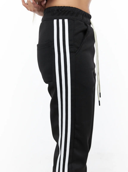Flared Striped Track Pants CU524 Black close-up side detail showing triple white stripe tape, side pocket, and cream drawstring at waist