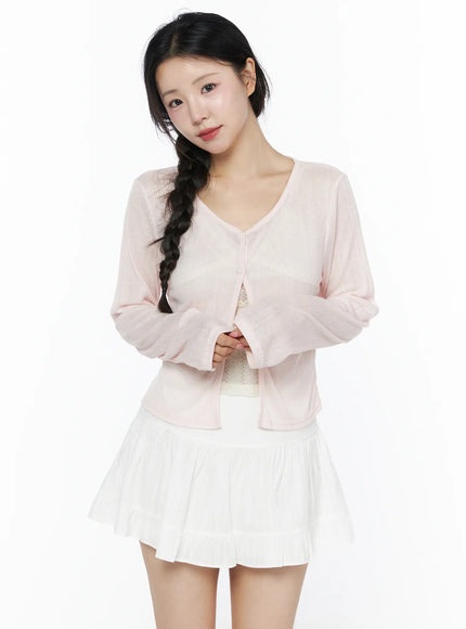 Flower Button Ribbed Cardigan IU525 Light pink front model shot hands clasped at waist showing V-neckline button front sheer ribbed knit and cropped silhouette over white pleated skirt