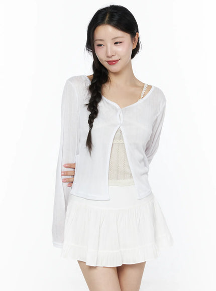 Flower Button Ribbed Cardigan IU525 White front model shot smiling showing sheer ribbed knit open-front cardigan with single button layered over cream lace camisole and white pleated skirt