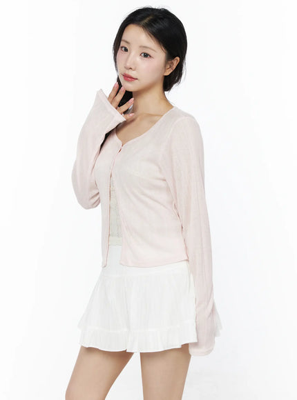 Flower Button Ribbed Cardigan IU525 Light pink three-quarter front model shot hand raised to chin showing open button front and sheer ribbed fabric over white skirt