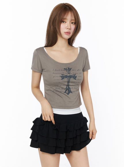 Gothic Cross Slim Fit Top IU525 front model shot in grey-brown color, scoop neck with dark cross and script print, layered over white inner, styled with black ruffle skirt