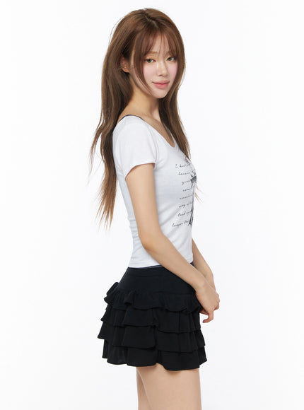 Gothic Cross Slim Fit Top IU525 White side model shot, smiling profile pose, cross and script print partially visible, slim stretchy crop fit, black tiered ruffle mini skirt
