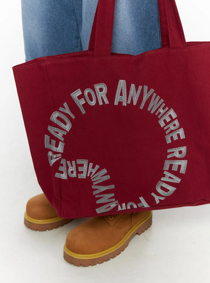 Graphic Canvas Tote Bag IL520 Dark red close-up front detail showing circular embroidered lettering on canvas fabric held at knee level