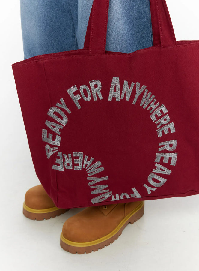 Graphic Canvas Tote Bag IL520 Dark red close-up front detail showing circular embroidered lettering on canvas fabric held at knee level