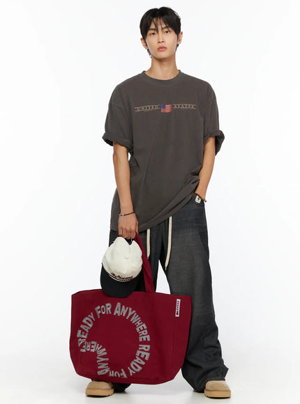 Graphic Canvas Tote Bag IL520 Dark red full front model shot male model holding bag at side styled with gray graphic tee dark jeans and boots