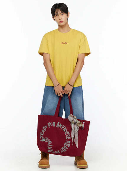 Graphic Canvas Tote Bag IL520 Dark red front full-body model shot male model carrying bag on shoulder wearing yellow tee wide-leg jeans and boots