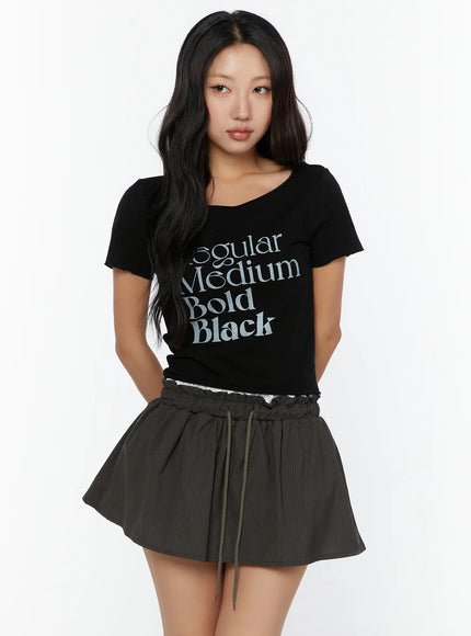 Graphic Cropped Tee CU526 Black front model shot with typography graphic print, scoop neckline, cropped fit, styled with dark mini skirt