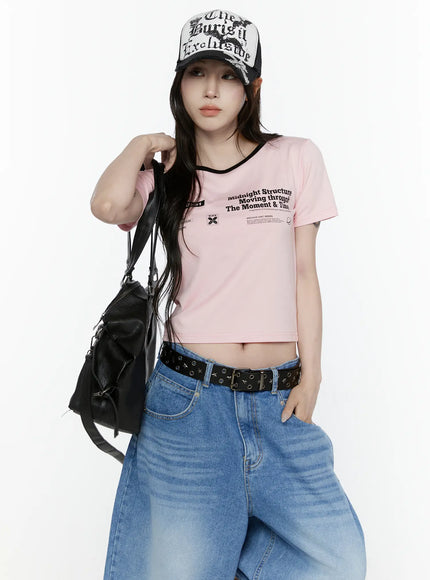 Graphic Cropped Tee IL521 Light pink front view on model with cap and black bag over shoulder, looking upward, cropped hem exposing midriff, styled with wide-leg jeans