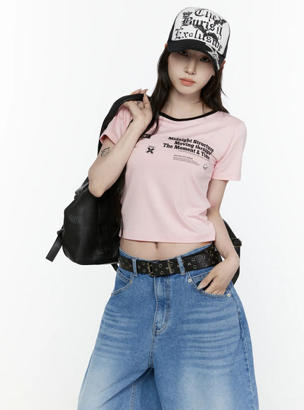 LEWKIN Graphic Cropped Tee IL521 Light pink S/M front view on model wearing cap and backpack, black ringer neckline, archive graphic print, styled with wide-leg jeans