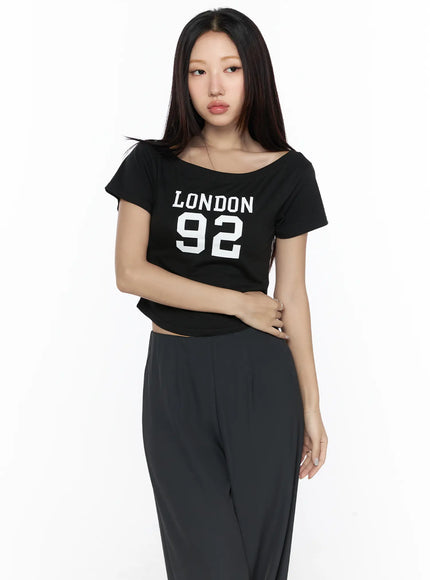 Graphic Off-Shoulder Crop Top CL524 Black front shot on model with arms crossed showing LONDON 92 print wide-neck crop fit with wide-leg trousers
