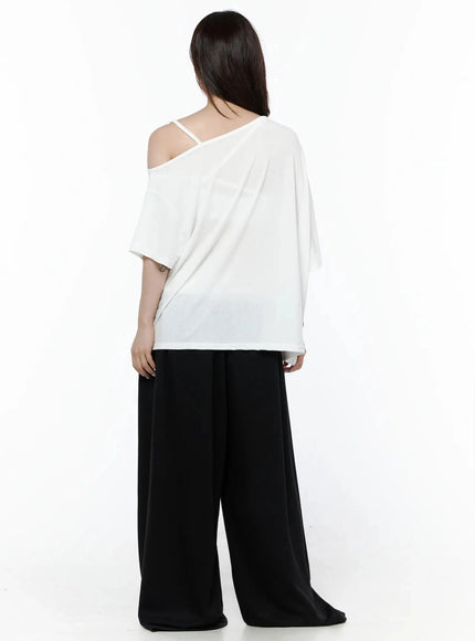 Graphic Off-Shoulder T-Shirt IL506 White back view on model, plain white oversized back, one-shoulder strap visible, soft drape fabric, styled with black wide-leg pants