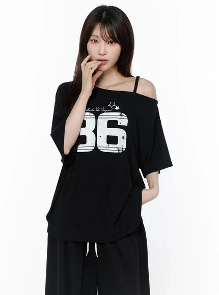 Graphic Off-Shoulder T-Shirt IL506 Black front view on model, oversized fit, white 86 distressed graphic print, one-shoulder strap detail, paired with black wide-leg trousers