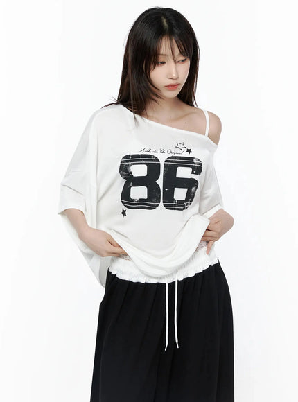 Graphic Off-Shoulder T-Shirt IL506 White front view on model lifting hem to show lace drawstring waistband detail, black 86 print, one-shoulder strap, black wide-leg pants