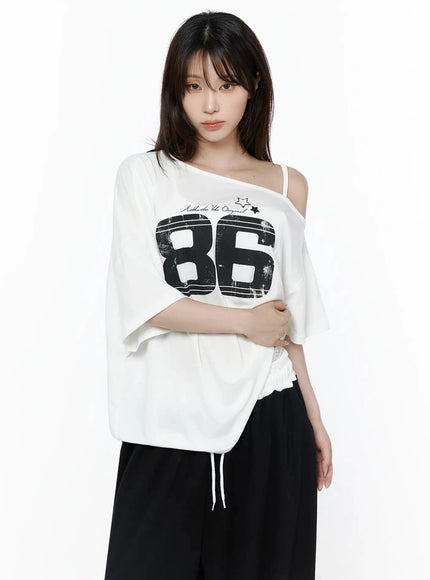 Graphic Off-Shoulder T-Shirt IL506 White front view on model with arm crossed at waist, black 86 distressed graphic, one-shoulder strap, lace hem drawstring, black trousers