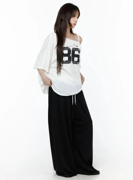 Graphic Off-Shoulder T-Shirt IL506 White side view on model, 3/4 angle showing 86 graphic, asymmetric one-shoulder strap, oversized drop-shoulder sleeve, black wide-leg trousers
