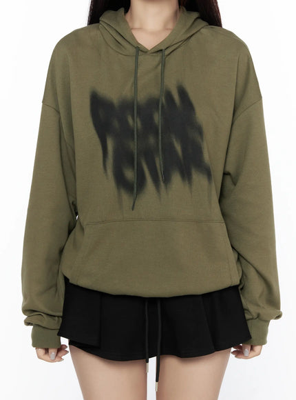 Graphic Oversized Hoodie CL524 Dark green close-up front detail on model showing blurred black graffiti-style chest graphic, drawstring hood, and ribbed cuffs