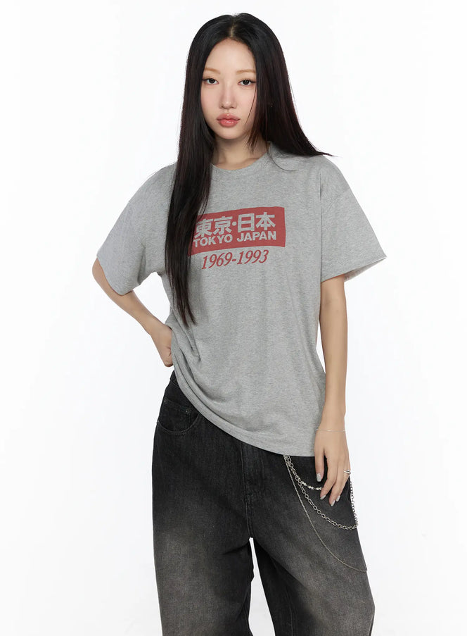 Graphic Oversized Tee CL527 Gray front shot on model hand on hip blue box Tokyo Japan 1969-1993 print styled with black jeans
