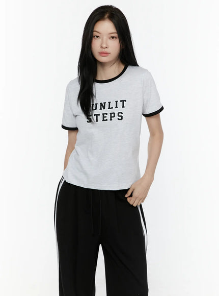 LEWKIN Graphic Ringer Tee IL506 Black front view on model wearing black track pants, light gray melange cotton tee with black ringer trim and SUNLIT STEPS varsity print