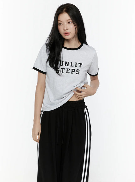 Graphic Ringer Tee IL506 Black three-quarter front view on model looking aside, hand gathering hem, light gray cotton tee with black ringer detail and stripe pants