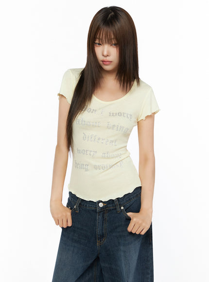 Graphic Scoop Neck Top IL523 Yellow half-body front shot on model looking down, hands in pockets, lettuce hem and rhinestone script detail highlighted