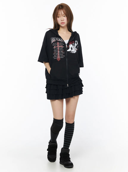 Graphic Short Sleeve Zip-Up Hoodie IU525 Black front view on model standing, LEEFOREN graphic print with red star motif, zip-up hoodie, styled with black ruffle mini skirt, knee socks