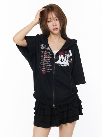 Graphic Short Sleeve Zip-Up Hoodie IU525 Black front view on model hand in hair casual pose, zip-up open slightly, red graphic print, worn over white tank with ruffle skirt