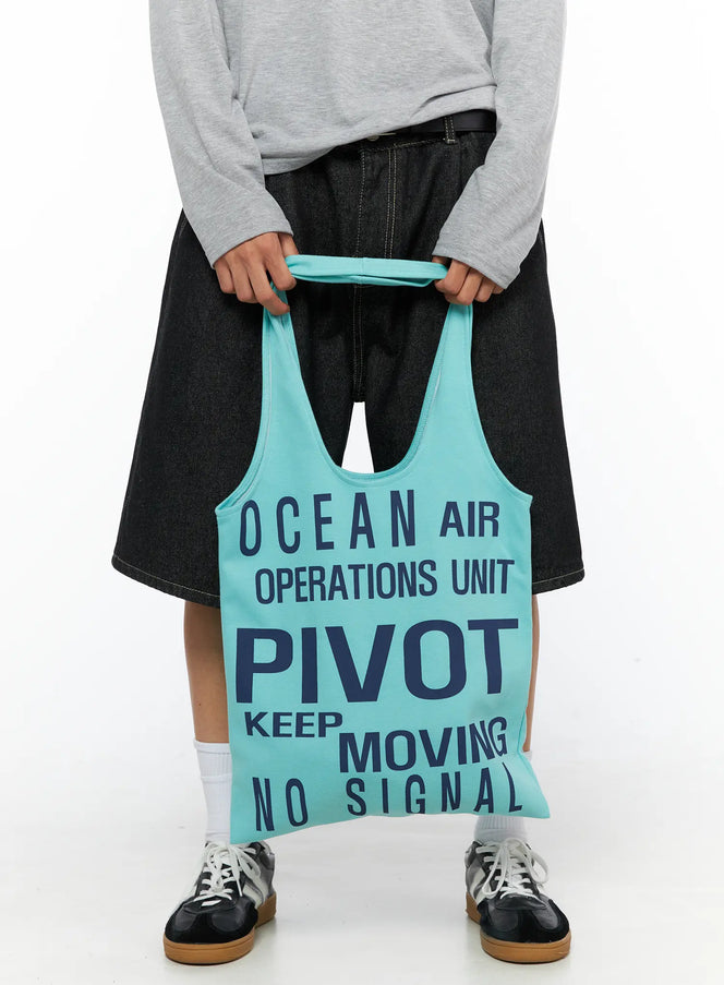 Graphic Tote Bag IL528 Mint front shot on model held with both hands at waist level navy PIVOT text print soft cotton fabric with denim shorts