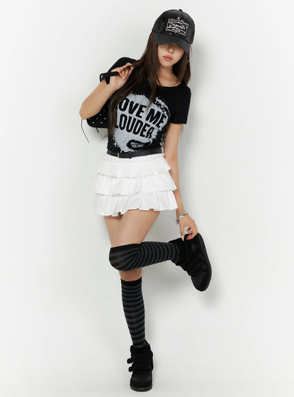 Graphic U-Neck Tee IL523 Black full-length front view on model with cap and bag accessory, playful pose lifting leg, LOVE ME LOUDER print, striped knee socks