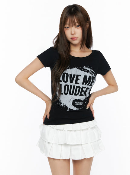 Graphic U-Neck Tee IL523 Black front view on model hands on hips, fitted short-sleeve tee with large LOVE ME LOUDER graphic, styled with white ruffle mini skirt