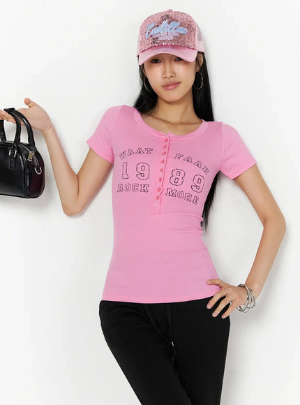 Henley Graphic Tee IL509 Pink front lifestyle shot model leaning on wall hand on hip holding black bag wearing pink sequin cap showing graphic print and button placket