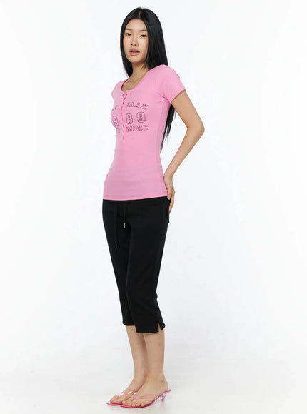 Henley Graphic Tee IL509 Pink angled front shot model turned slightly left showing ribbed texture button placket and WAAT FAAK 1989 graphic styled with black capri pants