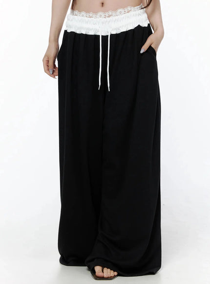 High-Waist Lace Wide Leg Sweatpants IL506 Black front model shot waist-down with hand in pocket showing lace waistband white drawstring and relaxed wide-leg fit