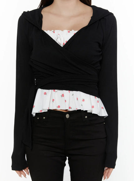 Hooded Wrap Crop Cardigan CL507 Black close-up front shot on model showing crossover wrap bodice, hooded neckline, white rose-print pleated ruffle hem insert detail