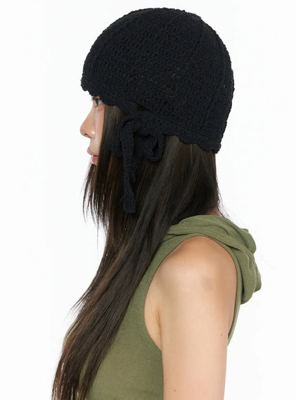 Knit Ribbon Beanie CL517 Black side profile on model in green tank top, showing rounded crochet silhouette and knotted ribbon tie hanging at ear