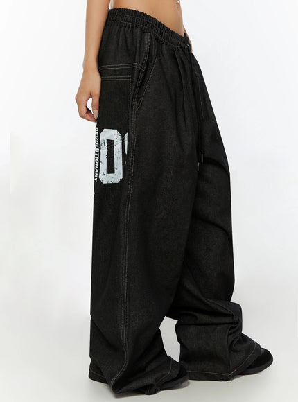 Kris Back Graphic Baggy Jeans IL524 Black side waist-down shot on model showing REVOLUTIONARY 08 white distressed graphic print on outer thigh and wide-leg hem