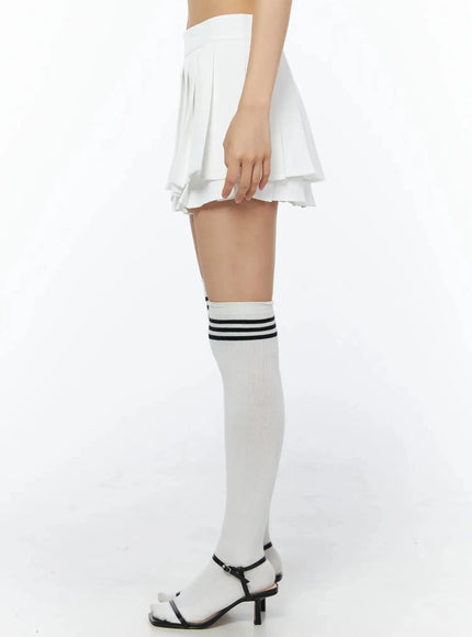Layered Pleated Skirt IL509 White side close-up on model showing single-leg view of layered pleated hem, over-the-knee white sock with black stripes, black sandal