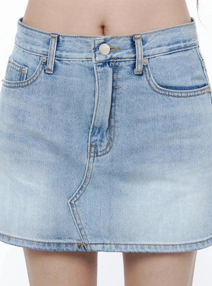Low-Rise Denim Mini Skirt IL502 Light blue close-up front detail showing silver button, belt loops, front pockets and diagonal seam stitching on model