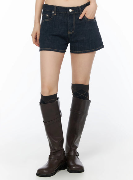 Low-Rise No-Fade Denim Shorts IU525 Dark blue front crop shot on model with hand on pocket, showing low-rise fit and gold contrast stitching with brown boots