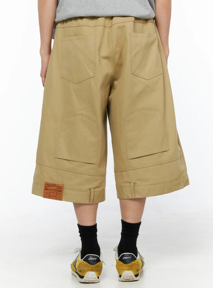 LEWKIN Men's Baggy Bermuda Shorts IL514 Beige back view on model wearing gray tee and yellow sneakers, wide-leg silhouette with cargo pockets and leather patch label