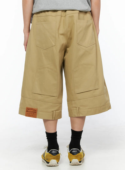 LEWKIN Men's Baggy Bermuda Shorts IL514 Beige back view on model wearing gray tee and yellow sneakers, wide-leg silhouette with cargo pockets and leather patch label