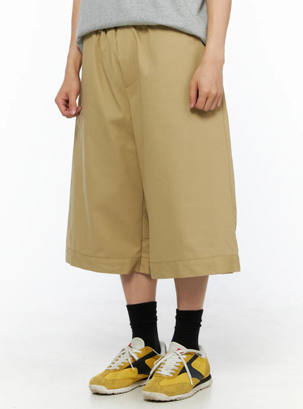 Men's Baggy Bermuda Shorts IL514 Beige front angled close-up on model showing side pocket seams, slightly asymmetric hem overlap, and cuffed bottom with yellow sneakers