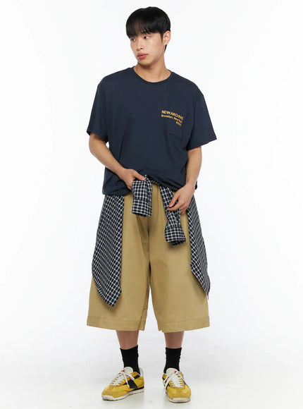 Men's Baggy Bermuda Shorts IL514 Beige front full-length view on model wearing navy graphic tee with plaid shirt tied at waist, wide-leg fit styled with yellow sneakers