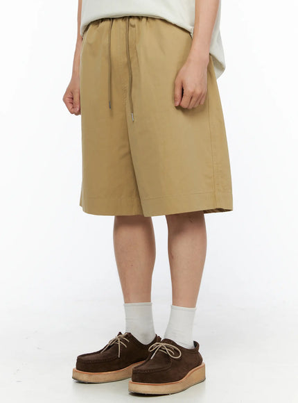 Men's Bermuda Shorts (Beige) IL514 beige angled front shot on male model showing drawstring waist elastic band and knee-length wide-leg fit with white socks