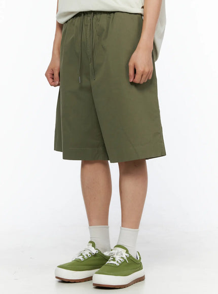 Men's Bermuda Shorts (Dark Green) IL514 Dark green side crop shot on model showing relaxed wide-leg cut, hem stitching, and side seam detail with green sneakers