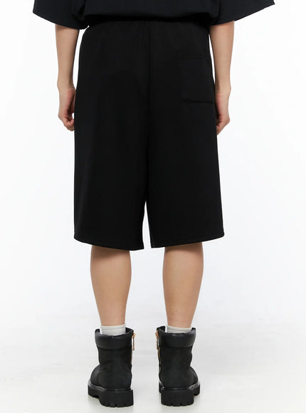 Men's Bermuda Sweat Shorts (Black) IL514 Black back shot on female model showing back patch pocket, center seam, elastic waistband, and wide-leg knee-length cut with black boots