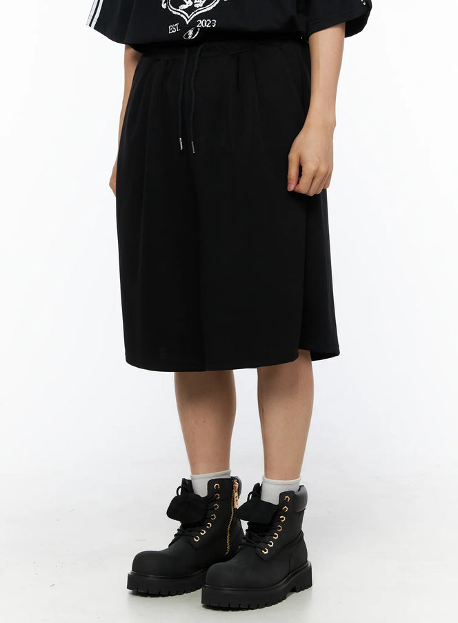 Men's Bermuda Sweat Shorts (Black) IL514 Black angled front shot on female model styled with black graphic tee and chunky boots, showing wide flowy hem and elastic waistband